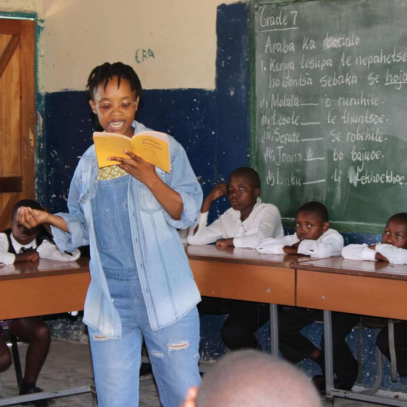 A young woman reads from a Sesotho literature book to a classroom of students